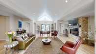 Located at 14505 Old Courthouse Way in Newport News, Virginia, this single-story leasing office offers a thoughtfully designed and inviting environment for residents and prospective tenants alike. The space is defined by a striking vaulted ceiling with a large triangular clerestory window at the entrance, flooding the interior with natural light and creating an immediate sense of openness and warmth upon arrival.

The main living area serves as a comfortable lounge, featuring light hardwood-style flooring throughout, recessed lighting, and a standout stacked-stone fireplace with a mounted television — providing a cozy and polished focal point for the room. Plush leather seating, including a sage green sofa and rich brown accent chairs, are arranged around a central woven area rug and a round wood coffee table, creating an inviting atmosphere for conversation and relaxation.

The reception area includes a sleek, modern desk with a warm wood-paneled front and dark countertop, positioned to offer views to both the entrance and an outdoor pool area. Open shelving and a secondary seating nook nearby provide additional functional space for meetings or consultations.

The kitchen is clean and contemporary, outfitted with white shaker cabinets, light stone countertops, stainless steel appliances, and a decorative green botanical wall feature. Open shelving adds a decorative yet practical touch, completing this well-equipped communal space.
