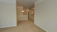 This single-floor residence showcases a thoughtfully designed open-concept layout that connects the living, dining, and kitchen areas in a cohesive and functional flow. The interior is finished with elegant crown molding that runs throughout, adding a refined architectural touch to the neutral, warm-toned walls. Plush wall-to-wall carpeting in the main living areas contributes to a comfortable and inviting atmosphere. A decorative column elegantly defines the transition between the living and dining spaces, lending a sense of structure without sacrificing the open feel of the floor plan. The dining area is highlighted by a classic pendant light fixture that draws the eye upward and adds a warm, ambient glow. Beyond the dining space, the kitchen features a raised bar-height counter, ideal for casual dining or entertaining, and is equipped with track lighting overhead. Wood-toned cabinetry adds warmth to the kitchen area. Generous baseboards complement the crown molding throughout, giving the space a polished, well-finished look. The neutral color palette and open layout make this residence highly adaptable, offering a clean and versatile canvas for a variety of interior styles.