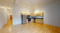 This single-floor unit offers a bright, modern living environment defined by light hardwood flooring that runs consistently throughout the space, creating a sense of warmth and continuity. The open kitchen is a standout feature, equipped with a full suite of stainless steel appliances including a refrigerator, electric range, built-in microwave, and dishwasher. Light maple cabinetry lines the walls, offering generous storage, while dark countertops provide a striking contrast. A track lighting system mounted along the ceiling ensures excellent illumination across the kitchen and adjoining living area. The soaring ceiling height adds an airy, loft-like quality to the space. A long, well-lit hallway connects the main living areas to the private rooms, maintaining a clean and organized flow throughout. The bathroom features the same warm hardwood flooring, matching maple vanity cabinetry with a white countertop and undermount sink, a wall-mounted mirror, and a walk-in shower with a white surround. The overall design is clean and contemporary, with white walls and minimal ornamentation allowing the architectural elements and finishes to take center stage, resulting in a bright, functional, and inviting living environment.