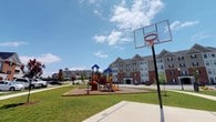 The Spring Creek community outdoor amenity space offers a well-appointed recreational area designed to serve residents of all ages. The centerpiece of the space includes a full-size basketball hoop with a clear backboard mounted on a paved half-court, providing a dedicated surface for play and activity. Adjacent to the court, a vibrant, multi-feature playground structure with slides, climbing elements, and colorful canopies sits atop a safety-surfaced base, making it an inviting space for younger residents. The surrounding landscape is characterized by expansive, neatly maintained green lawns that create an open, airy atmosphere and offer ample open space for outdoor recreation. Mature trees and ornamental plantings are scattered throughout, adding shade and visual interest to the grounds. In the background, the multi-story residential buildings feature classic brick and neutral-toned exteriors with symmetrical window arrangements, contributing to an orderly and cohesive neighborhood aesthetic. The entire outdoor area benefits from generous natural light and open skies, reinforcing a welcoming and active community environment that prioritizes outdoor living and social engagement for all residents.