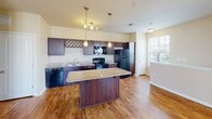 This single-floor residence offers a thoughtfully designed layout that balances open living spaces with comfortable private quarters. The kitchen is a standout feature, showcasing rich dark cabinetry with a decorative wine rack accent, granite countertops, a center island with pendant lighting overhead, and a full suite of black appliances including a dishwasher, range, and refrigerator. Warm hardwood-style flooring runs throughout the kitchen and extends seamlessly into the open-concept dining and living area. The living space is bright and airy, benefiting from multiple windows fitted with horizontal blinds, a ceiling fan, and an elegant arched architectural detail that frames the transition between areas. The bedrooms are carpeted for comfort, each featuring ceiling fans and windows that allow natural light to filter in. The primary bedroom connects to a bathroom equipped with a vanity featuring dark cabinetry and granite-style countertops, a large mirror, and a combination tub and shower. A secondary bathroom mirrors this practical design. Throughout the residence, neutral warm-toned walls, clean white trim, and consistent finishes create a cohesive and inviting atmosphere well-suited for everyday living.