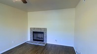 This single-story space presents a clean and functional layout highlighted by several notable design elements. The living area centers around a fireplace framed by a tile surround and raised tile hearth, adding a classic focal point to the room. The tile work features a neutral stone-like pattern that complements the overall color scheme.

Dark hardwood flooring runs throughout the space, providing warmth and contrast against the light, neutral-toned walls. The smooth, white ceiling enhances the sense of openness, while a ceiling fan provides practical climate control. Crisp white baseboards offer a clean finishing detail throughout.

The walls are painted in a soft, warm off-white tone, creating a bright and welcoming atmosphere that serves as a versatile backdrop for a variety of interior design styles. The space feels open and uncluttered, offering flexibility for furniture arrangement and personalization.

Overall, the space strikes a balance between simplicity and comfort, with the fireplace serving as its defining architectural feature. The combination of dark flooring, neutral walls, and classic tile detailing creates a cohesive and inviting environment.