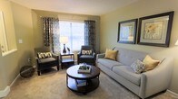 This two-floor space offers a comfortable and well-appointed living area as seen in the main gathering room. The living space is finished with soft, neutral-toned carpeting that adds warmth underfoot, while light sage-green walls create a calm and inviting atmosphere throughout. A large window dressed with bold geometric-patterned curtains and horizontal blinds allows generous natural light to fill the room, enhancing its open and airy feel. The seating arrangement is thoughtfully organized around a dark wood round coffee table, which serves as a central anchor for the space. Coordinating upholstered armchairs and a cushioned sofa in muted grey and cream tones provide ample seating, complemented by decorative throw pillows in gold and neutral hues. A side table with a table lamp adds a functional yet stylish accent. Framed wall art contributes a finished, polished look to the room. The overall ambiance is warm, neutral, and livable, with a cohesive design aesthetic that balances comfort and style. The two-floor layout suggests additional living or sleeping areas beyond what is visible, offering versatile use of space.