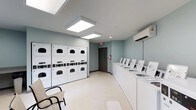 This communal laundry facility offers a clean, functional, and well-organized environment designed to accommodate multiple users efficiently. The room is finished with soft blue-gray walls and light-toned flooring, creating a calm and tidy atmosphere throughout.

Along one wall, a bank of stacked commercial-style dryers provides generous drying capacity, each unit equipped with coin-operated controls. Opposite, a row of top-loading washing machines lines the adjacent wall, offering ample washing stations for simultaneous use. The layout is thoughtfully arranged to allow ease of movement between machines.

Overhead, large flush-panel light fixtures illuminate the space evenly, ensuring a bright and comfortable environment. A wall-mounted mini-split air conditioning unit maintains climate control, keeping the room cool and pleasant during use. A fire extinguisher is visibly mounted near the exit door, reflecting attention to safety standards.

A seating area with chairs is positioned near the entrance, providing a convenient spot to wait while laundry cycles complete. The single-floor layout, wide-open floor plan, and clearly marked exit contribute to an accessible and user-friendly space. Overall, this laundry room is a practical, well-maintained facility built to serve the needs of a larger residential or hospitality setting.