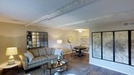 This single-floor space offers an open-concept layout that connects the living and dining areas in a cohesive, functional design. The living area is anchored by a comfortable striped sofa, a glass-topped coffee table, and decorative accents including a large black-framed grid mirror that adds depth and visual interest to the room. Exposed ceiling beams with a whitewashed finish lend a rustic yet refined character to the space, complementing the warm hardwood-style flooring that runs throughout.

A seamless transition leads into the dining area, where a round dark-wood table is surrounded by classic Windsor-style chairs, creating an intimate setting for meals. A vintage-style wall clock and a framed map serve as tasteful wall décor, reinforcing a timeless, collected aesthetic. Soft, neutral wall tones throughout the space enhance its welcoming ambiance, while a combination of overhead and lamp lighting ensures a warm, well-lit environment. A second framed map panel near the living area adds a decorative accent that ties the two zones together visually.
