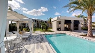 This beautifully designed outdoor amenity space embodies a resort-style atmosphere, centered around a pristine swimming pool with crystal-clear turquoise water. The pool deck is finished with elegant paver stones in a multi-toned pattern, providing a sophisticated and durable surface that complements the overall aesthetic. A private cabana with flowing white curtains and comfortable lounge seating offers a shaded retreat, ideal for relaxing in style. Adjacent to the pool, a sleek poolside structure with a dark awning serves as a covered service or amenity station, adding both function and visual contrast to the space. Lush tropical landscaping, including tall swaying palm trees and manicured greenery, frames the area and enhances the warm, resort-like ambiance. The surrounding multi-story residential building provides an elegant architectural backdrop, while the open sky above ensures the space is bathed in abundant natural light. The thoughtful layout balances open sun-soaked areas with shaded retreats, catering to a variety of outdoor leisure preferences. Overall, this space delivers a polished, high-end outdoor experience reminiscent of a luxury resort destination.