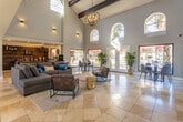 Located at 535 S Mesa Hills Dr in El Paso, TX, this single-floor space serves as an inviting communal lounge or clubhouse with a striking architectural presence. The interior is immediately defined by its dramatic vaulted ceilings adorned with exposed dark wood beams and a statement chandelier, creating a grand sense of volume and openness. Large arched clerestory windows line the upper walls, flooding the space with natural light alongside oversized picture windows at ground level that frame views of the surrounding palm-lined outdoor grounds and pool area. The main lounge area features a generously sized sectional sofa and tufted leather accent chairs arranged around a vibrant area rug and natural wood stump side tables, offering a comfortable and eclectic gathering space. A built-in bar area with a reclaimed wood backsplash and pendant lighting provides a stylish focal point, ideal for social gatherings. A dedicated dining nook with a round wood table and modern upholstered chairs sits beside floor-to-ceiling windows, offering serene outdoor views. French doors open directly to the exterior, seamlessly connecting the indoor and outdoor environments. Large-format tile flooring throughout ties the space together with a clean, contemporary aesthetic.
