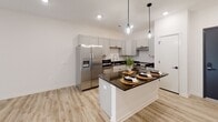 This single-floor space is designed with a clean, contemporary aesthetic that emphasizes an open and functional layout. The kitchen serves as a central highlight, featuring gray shaker-style cabinetry that extends to the upper walls, dark granite countertops, and a full suite of stainless steel appliances including a side-by-side refrigerator with water dispenser, a freestanding range, and a built-in microwave. A generously sized kitchen island with matching dark countertops provides additional prep space and casual seating for multiple guests, accented above by stylish globe pendant lights that add a modern touch.

The kitchen flows seamlessly into the open living area, which is finished with light wood-style plank flooring that runs throughout the main living spaces, creating a cohesive and airy feel. Large sliding glass doors flood the living area with natural light and provide access to an outdoor space. The bedroom features soft carpeting, a ceiling fan, and a window with a roller shade. The bathroom is finished with the same light flooring and includes an accessible shower area with black hardware accents. Throughout the space, crisp white walls and recessed lighting contribute to a bright, minimalist ambiance.