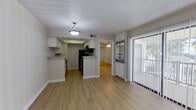 This single-story unit is finished consistently throughout with light hardwood-style flooring and neutral gray-toned walls, creating a cohesive and clean aesthetic from room to room. The living area is an open, well-proportioned space anchored by a sliding glass door fitted with vertical blinds, offering direct access to an outdoor balcony and views of surrounding greenery. Natural light filters into the room, complementing the bright, airy feel of the interior.

The galley-style kitchen is efficiently laid out with white cabinetry lining both walls, providing ample upper and lower storage. Light granite-style countertops contrast neatly with the black appliances, which include a refrigerator, electric range, microwave, and dishwasher. A built-in sink with a single basin is positioned along one wall, and a recessed ceiling detail adds subtle architectural interest overhead.

The bedroom is a calm, comfortable space featuring a ceiling fan with integrated lighting and a second sliding glass door with vertical blinds, allowing for additional natural light and outdoor access. Adjacent to the bedroom, the bathroom includes a vanity with white cabinetry and a mirror, maintaining the clean, consistent design language found throughout the unit. Textured ceilings are present throughout, adding a layer of traditional detail to the otherwise modern, streamlined finishes.