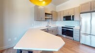 Located at 126 Langdon Street in Madison, Wisconsin, this thoughtfully designed 450-square-foot studio apartment offers a functional and modern living experience across a single floor. The kitchen is a standout feature, boasting gray shaker-style cabinetry, white quartz countertops, a classic white subway tile backsplash, and a full suite of stainless steel appliances including a refrigerator, range, microwave, and dishwasher. Open wood floating shelves add a contemporary touch, while a large angled peninsula provides additional counter space and doubles as a dining area with bar seating. Light hardwood-style flooring runs throughout the unit, lending warmth and continuity to the open layout. A bold yellow pendant light above the kitchen peninsula adds a pop of color and character. The bathroom features a clean, minimalist design with a white quartz vanity top, an undermount sink with chrome fixtures, gray cabinetry with chrome hardware, and a circular black-framed mirror. A dedicated laundry area houses a stacked Whirlpool front-loading washer and dryer alongside a wire shelving unit, maximizing convenience within the compact footprint. The doll's-eye view reveals a bright living area with a window, completing this well-appointed urban residence.