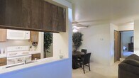 This single-floor residence offers a thoughtfully designed open-concept layout that connects the kitchen, dining, and living areas in a seamless flow. The kitchen features warm wood cabinetry, white appliances including a range and microwave, and a light-colored countertop pass-through that opens directly to the adjacent dining space — a practical design detail that enhances both functionality and visual connectivity between rooms. A dark wood overhead cabinet accent above the pass-through adds a bold, contrasting design element to the otherwise neutral color palette.

The dining area accommodates a round table with dark chairs and is anchored by a ceiling fan with a light fixture, which provides both air circulation and ambient lighting. The space is finished with plush wall-to-wall carpet in a neutral tone, contributing to a warm and comfortable atmosphere throughout. Light-colored walls keep the interior feeling bright and open. A hallway visible in the background suggests additional rooms beyond the main living areas. The overall ambiance is cozy and practical, with a clean, unpretentious aesthetic suited for everyday living.