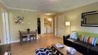 This single-floor residence offers a functional and comfortable layout designed to maximize everyday living. The open-concept living and dining area serves as the heart of the space, featuring warm wood-look flooring that runs throughout, adding a cohesive and inviting feel. Crown molding along the ceiling adds a subtle touch of elegance, while a textured ceiling and recessed ventilation contribute to the overall practical design. The living area provides ample room for seating and relaxation, complemented by a stylish floor lamp that offers warm ambient lighting. Adjacent to the living space, the dining area accommodates a modest dining table and chairs, making it well-suited for casual meals. A standout feature of this unit is the in-unit laundry area, conveniently located off the main living space and housing a full-size washer and dryer side by side — a practical amenity that adds significant convenience. A separate door leads to additional spaces within the unit, suggesting a thoughtful division between living and private areas. The warm, neutral wall tones throughout create a welcoming atmosphere, making this a cozy and well-appointed single-floor living space.