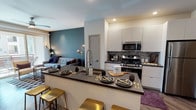 This single-floor residence showcases a thoughtfully designed open-concept layout that seamlessly connects the kitchen, dining, and living areas. The kitchen is a standout feature, outfitted with stainless steel appliances including a gas range, over-the-range microwave, and a full-size refrigerator. Sleek white cabinetry with dark countertops provides ample storage and a striking contrast, while a textured stone tile backsplash adds visual depth and character. A peninsula breakfast bar with a dark countertop and gold bar stools serves as both a functional dining space and a social hub, anchored by a modern pull-down faucet. Recessed lighting throughout ensures the space is well-lit and inviting. Rich wood-look flooring runs throughout the common areas, lending warmth and continuity. The living area, visible from the kitchen, features a bold teal accent wall that adds a pop of color against neutral tones, complemented by stylish wall sconces. A dedicated laundry area with a stacked washer and dryer unit is conveniently located just off the kitchen. The overall ambiance is contemporary and vibrant, balancing modern finishes with warm, welcoming design elements.
