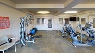 Located at 3304 E 44th Ave in Spokane, Washington, this single-floor fitness center is a well-equipped exercise facility designed to support a full range of workout routines. The space features a thoughtfully arranged selection of cardio equipment, including treadmills, elliptical trainers, and stationary bikes, providing ample options for aerobic exercise. A multi-station strength machine, weight bench, and stability balls round out the strength and flexibility offerings.

The room is finished with durable, patterned carpet tiles and warm neutral-toned walls, creating a functional and welcoming environment. A large wall-mounted mirror extends along one side, enhancing the sense of space and allowing users to monitor their form during workouts. Recessed ceiling panels with flush lighting provide consistent, even illumination throughout the room.

A wall-mounted flat-screen television is positioned for easy viewing from the cardio area. Hand sanitizer dispensers are conveniently placed near the entrance. The fitness center benefits from abundant natural light, courtesy of multiple windows and a full glass sliding door fitted with vertical blinds that opens directly onto an outdoor balcony. The balcony overlooks the surrounding Spokane landscape, offering a refreshing outdoor connection to complement the indoor workout experience.