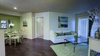 This single-floor space offers a clean, well-organized layout designed for comfortable everyday living. The living area serves as the heart of the home, featuring rich dark hardwood flooring that contrasts beautifully with the light, neutral-toned walls. A textured ceiling adds subtle character to the room, while contemporary artwork adorns the walls, contributing to an inviting and polished ambiance. The living space is furnished with a plush white sofa accented with patterned throw pillows, a glass-top coffee table with wrought iron detailing, and a light green area rug that ties the room together. A classic white paneled door leads to an adjacent room, suggesting a functional and well-connected floor plan. The space benefits from ample ambient lighting, creating a bright and airy atmosphere throughout. The overall design aesthetic is cohesive and understated, blending practical furnishings with decorative touches such as framed canvas paintings and potted greenery. The combination of warm flooring, soft wall tones, and curated décor makes this a welcoming and livable space suitable for a variety of lifestyles.