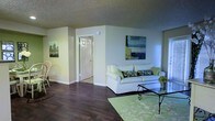 This single-floor space offers a clean, well-organized layout designed for comfortable everyday living. The living area serves as the heart of the home, featuring rich dark hardwood flooring that contrasts beautifully with the light, neutral-toned walls. A textured ceiling adds subtle character to the room, while contemporary artwork adorns the walls, contributing to an inviting and polished ambiance. The living space is furnished with a plush white sofa accented with patterned throw pillows, a glass-top coffee table with wrought iron detailing, and a light green area rug that ties the room together. A classic white paneled door leads to an adjacent room, suggesting a functional and well-connected floor plan. The space benefits from ample ambient lighting, creating a bright and airy atmosphere throughout. The overall design aesthetic is cohesive and understated, blending practical furnishings with decorative touches such as framed canvas paintings and potted greenery. The combination of warm flooring, soft wall tones, and curated décor makes this a welcoming and livable space suitable for a variety of lifestyles.