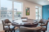 Located at 6604 Odana Rd in Madison, WI, this amenity space spans two floors and serves as a dynamic recreational hub within a modern multi-story residential building. The interior is anchored by a stylish game room featuring a full-size billiards table set beneath a contemporary linear pendant light, with a bold teal accent wall adding a vibrant focal point. Patterned carpeting and curated wall art contribute to an inviting, lively atmosphere. Adjacent to the billiards area is a comfortable lounge with a large wall-mounted television, leather sofa, upholstered seating, and a round coffee table — ideal for relaxing or socializing. A built-in kitchenette with light wood cabinetry, a sink, and a beverage refrigerator provides added convenience. The space connects directly to a generous outdoor courtyard enclosed by the surrounding building, finished with large-format white tile. The courtyard is outfitted with ample seating arrangements, dining tables, a fire pit, and a built-in grilling and prep station, creating a well-equipped outdoor gathering area. Upper-level balconies overlook the courtyard, while an exterior terrace offers open views of the surrounding neighborhood.