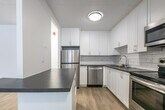 Located at 1001 Talwood Dr in Peterborough, Ontario, this 2-bedroom, 2-bathroom unit offers a clean, modern aesthetic throughout its thoughtfully designed single-floor layout. Light wood vinyl flooring flows seamlessly through the living spaces, complemented by crisp white walls and consistent flush-mount lighting.

The kitchen is a standout feature, showcasing white shaker-style cabinetry, a white subway tile backsplash, dark countertops, and a full suite of stainless steel appliances including a range, over-the-range microwave, dishwasher, and refrigerator. A peninsula counter connects the kitchen to the combined dining and living area, which benefits from a large floor-to-ceiling window dressed with vertical blinds, flooding the space with natural light.

Both bedrooms are well-sized, with the generously proportioned primary bedroom offering ample space. Each bedroom features light wood flooring, white walls, and baseboard heating. One bedroom includes dual windows with blinds, providing additional natural light.

The two bathrooms are finished with modern fixtures. One features a dark wood-toned vanity with a stone-look countertop and chrome hardware, while the other includes a bathtub/shower combination clad in classic white subway tile. A hallway with dedicated closet space completes the unit's practical layout.