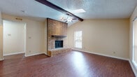 This single-story home presents a thoughtfully arranged open layout connecting the main living areas with functional flow throughout. The living room serves as the focal point of the home, showcasing vaulted ceilings accented by a dark exposed beam and a full-height brick fireplace with a raised brick hearth. A ceiling fan is centrally mounted, and a skylight adds natural illumination to the vaulted ceiling space. Rich, dark wood-look flooring extends across the living and bedroom areas, complementing the neutral wall tones throughout.

An open sightline from the living room leads toward the kitchen, which features warm honey-toned cabinetry, granite-look countertops, tile flooring, and a full suite of stainless steel appliances including a refrigerator, range, and over-the-range microwave. The bathroom is well-appointed with a dual-sink vanity topped with stone-look counters, warm wood cabinetry, tile flooring, and a tub/shower combination enclosed by sliding glass doors. Additional rooms feature the same wood-look flooring and ceiling fans. A secondary entry door with adjacent windows brings in natural light near what appears to be a utility or transitional space.