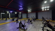 Located at 697 Bronx River Rd in Yonkers, NY, this single-floor fitness studio is purpose-built for indoor cycling and group fitness classes. The space is defined by its bold, high-energy aesthetic: matte black walls and ceiling contrast sharply with vivid yellow accent trim running along the base of the walls, while exposed red overhead pipes add an industrial character to the overall design.

The main cycling area is generously sized, accommodating a full fleet of stationary spin bikes arranged in rows across light wood-style vinyl flooring, which provides both durability and a clean, modern look. Full-length mirrors line one wall, enhancing the sense of space and allowing participants to monitor their form during workouts. Track lighting fixtures mounted to the dark ceiling deliver focused illumination throughout the room, complemented by atmospheric purple accent lighting that contributes to an energized, studio-like ambiance.

Structural columns are integrated cleanly into the layout, and a wall-mounted HVAC unit ensures climate control during high-intensity sessions. Along one wall, shelving units store yoga mats, foam blocks, resistance bands, and other fitness accessories, indicating the studio's versatility beyond cycling. A punching bag is also visible, suggesting additional training options. The space is well-organized and designed to support a motivating group fitness environment.