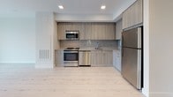 Located at 205 Revere Beach Pkwy in Revere, Massachusetts, this modern single-floor unit offers a thoughtfully designed open layout with a clean, contemporary aesthetic throughout. Light-toned hardwood flooring flows seamlessly across the main living areas, complementing the crisp white walls and creating a bright, airy atmosphere.

The open living area is generously proportioned and bathed in natural light from large floor-to-ceiling windows, which frame views of the surrounding Revere neighborhood. The kitchen is fully equipped with stainless steel appliances, including a gas range, over-the-range microwave, dishwasher, and refrigerator. Light wood cabinetry extends from floor to ceiling, paired with a chevron-patterned tile backsplash and light quartz countertops, delivering a cohesive and modern finish.

The bathroom features a floating wood-toned vanity with a white countertop, a backlit mirror, and large-format white tile flooring, contributing to a clean and minimalist design. Additional storage is provided through a spacious walk-in closet outfitted with wire shelving and carpeted flooring, offering ample organizational capacity. Recessed lighting throughout the unit ensures consistent illumination, while the overall layout maximizes functionality and flow within the space.