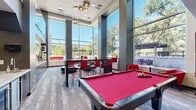 Located at 839 16th St SW in Rochester, Minnesota, this clubroom is a well-appointed community gathering space designed for socializing and recreation. The room impresses immediately with its soaring double-height ceilings and expansive floor-to-ceiling windows that flood the interior with natural light and offer views of the surrounding outdoor terrace and mature trees beyond.

The space is anchored by a red felt pool table positioned centrally on patterned carpet flooring, serving as the focal point for recreation. A built-in wet bar with granite countertops and dark cabinetry lines one wall, providing a convenient service area for gatherings. On the opposite end, a dark wood-paneled media wall houses a flat-screen television, flanked by decorative accessories and complemented by a comfortable sectional sofa and a round coffee table — ideal for relaxed viewing.

High-top dining tables with bold red chairs add a lively, modern energy to the room's palette of grays and reds. Sculptural globe pendant lights hang from the ceiling, adding an elegant touch. A glass-railed mezzanine level is visible above, contributing to the open, airy volume of the space. Direct access to an outdoor terrace further extends the usability of this dynamic clubroom.