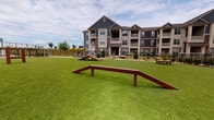 Located at 2024 Barr Rd in Kannapolis, North Carolina, this outdoor amenity space is a dedicated dog park designed with both pets and their owners in mind. The expansive area is covered in lush artificial turf, providing a clean, low-maintenance surface ideal for year-round use. Multiple wooden agility structures, including ramps and hurdles, are distributed throughout the space, offering engaging physical activity for dogs of various sizes. A standout feature is the timber-framed pavilion, constructed with natural wood posts and an open-beam ceiling, which provides a shaded retreat for residents. Beneath the pavilion, a concrete patio is furnished with Adirondack-style chairs and a side table, creating a comfortable seating area where owners can relax while supervising their pets. Decorative rock landscaping borders portions of the perimeter, adding a natural, polished aesthetic. The entire area is enclosed by a sturdy black metal fence, ensuring the safety and security of all animals. A pet waste station is also present, promoting cleanliness throughout the space. The park is set against the backdrop of a multi-story residential building featuring a combination of board-and-batten siding, stone accents, and private balconies, contributing to the overall well-maintained and community-oriented character of the property.
