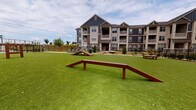 Located at 2024 Barr Rd in Kannapolis, North Carolina, this outdoor amenity space is a dedicated dog park designed with both pets and their owners in mind. The expansive area is covered in lush artificial turf, providing a clean, low-maintenance surface ideal for year-round use. Multiple wooden agility structures, including ramps and hurdles, are distributed throughout the space, offering engaging physical activity for dogs of various sizes. A standout feature is the timber-framed pavilion, constructed with natural wood posts and an open-beam ceiling, which provides a shaded retreat for residents. Beneath the pavilion, a concrete patio is furnished with Adirondack-style chairs and a side table, creating a comfortable seating area where owners can relax while supervising their pets. Decorative rock landscaping borders portions of the perimeter, adding a natural, polished aesthetic. The entire area is enclosed by a sturdy black metal fence, ensuring the safety and security of all animals. A pet waste station is also present, promoting cleanliness throughout the space. The park is set against the backdrop of a multi-story residential building featuring a combination of board-and-batten siding, stone accents, and private balconies, contributing to the overall well-maintained and community-oriented character of the property.