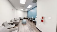Located at 405 W Redwood St in Baltimore, MD, this thoughtfully designed commercial lounge and breakroom space offers a bright, contemporary environment well-suited for collaborative gatherings or relaxed breaks. The single-level layout features wood-look luxury vinyl flooring throughout, providing a warm contrast to the crisp white walls and a bold teal accent wall that anchors the seating area at the rear.

Overhead, a drop ceiling with large recessed panel lighting ensures consistent, even illumination across the entire space. Seating is diverse and flexible, including modern grey upholstered lounge chairs with attached side tables, a round white marble-top table with molded chairs, a bar-height counter along the teal accent wall with stools, and a pair of light blue cushioned ottomans at the center for casual use.

A dedicated kitchenette alcove is finished with white subway tile backsplash and dark espresso cabinetry, housing a beverage station, sink, and undercounter refrigerator — ideal for daily refreshment needs. A glazed door with a metal frame provides access to an adjacent area, while a wall-mounted fire extinguisher and safety equipment reflect code-compliant, professional standards throughout. The overall ambiance is clean, functional, and inviting.