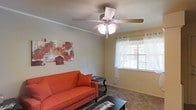 This single-floor space presents a welcoming living room as its central feature. The room is finished with neutral beige walls that create a warm, inviting atmosphere, complemented by carpeted flooring that adds comfort underfoot. A five-blade ceiling fan with an integrated light fixture is centrally mounted, providing both ventilation and ambient lighting.

Natural light filters into the space through a window dressed with horizontal blinds and sheer white curtains on a silver ring rod, offering both privacy and a soft glow. Crown molding runs along the upper perimeter of the walls, adding a subtle architectural detail that refines the overall look of the room.

The living area is furnished with a vibrant orange sofa accented by a matching throw pillow, positioned alongside a dark wood side table and a black coffee table. Abstract floral wall art in red and orange tones adds a pop of color and personality to the space. The overall ambiance is casual yet thoughtfully appointed, making it a functional and comfortable everyday living area.