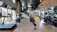 Located in Miramar, FL, this fitness center offers a well-appointed workout environment designed to support a full range of exercise routines. The space features an open, airy layout accentuated by soaring ceilings and abundant natural light streaming through large windows, providing views of the surrounding outdoor greenery. The interior design blends industrial and modern aesthetics, highlighted by exposed brick walls, a dark ceiling with recessed lighting, and warm wood-look flooring throughout.

The cardio zone is equipped with treadmills, elliptical machines, and a stair climber, each positioned near windows to allow for a pleasant outdoor view during workouts. Wall-mounted flat-screen televisions are conveniently placed throughout the space for entertainment. The strength training area houses a Life Fitness cable crossover machine, a leg press, and a dedicated free weights section complete with a dumbbell rack and floor-to-ceiling mirrors that visually expand the space.

A metal-railed staircase with open treads connects the main floor to an upper mezzanine level, adding a dynamic architectural element to the space. Decorative athletic-themed wall murals and medicine balls mounted near the ceiling contribute to the motivating ambiance. Glass partitions and large entry doors with exterior access complete the functional and polished design of this comprehensive fitness facility.