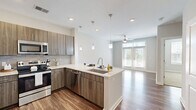 Located at 1055 Hatches Pond Ln in Cary, NC, this single-floor residence offers a thoughtfully designed layout that maximizes both functionality and comfort. The open-concept living and kitchen area is anchored by warm wood-look flooring and bathed in natural light from large windows and glass doors, creating a bright and welcoming ambiance throughout.

The kitchen is a standout feature, showcasing wood-toned cabinetry with sleek hardware, light granite countertops, a tile backsplash, and a full suite of stainless steel appliances including a refrigerator, range, microwave, and dishwasher. A peninsula with pendant lighting and an undermount sink provides additional prep space and connects seamlessly to the living area, which is finished with a ceiling fan overhead.

The spacious primary bedroom offers plush wall-to-wall carpeting, a ceiling fan with light, and a well-sized window with blinds that allows for natural light while maintaining privacy. The room includes closet access and is designed to comfortably accommodate a full bedroom suite. Additional storage and utility space are visible throughout the layout, including what appears to be an in-unit laundry area. Neutral tones and clean lines throughout give this residence a modern, move-in-ready feel.