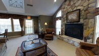 This inviting single-floor space exudes classic charm and warmth through its thoughtfully curated design elements. The room's most commanding feature is its dramatic floor-to-ceiling natural stone fireplace, which anchors the space and serves as a stunning focal point. Complementing this is a soaring vaulted ceiling that enhances the sense of openness and grandeur throughout the room. Natural light pours in generously through multiple large windows, including a graceful arched window flanking the fireplace, as well as a glass-paneled door that offers views to the outdoors. The interior is finished in deep, earthy tones with olive-green walls that create a rich, welcoming ambiance. Comfortable seating, including a plush upholstered armchair with a matching ottoman, is arranged to encourage relaxation and conversation. Decorative touches such as an ornate area rug, a glass-top coffee table with a brass globe accent, valance-adorned window treatments, and framed wall art lend a refined, traditional aesthetic. Recessed lighting and a classic floor lamp provide layered illumination, ensuring the space remains warm and functional at any time of day.
