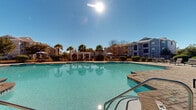 Located at 285 Park Ave in Pooler, Georgia, this outdoor amenity area showcases a resort-style environment designed for relaxation and recreation. At the heart of the space is a generously sized swimming pool with clear, blue-green water, accessible via wide entry steps with handrails. The pool is surrounded by an expansive paver deck crafted from richly textured brick-pattern stone, providing ample room for lounging and socializing. Rows of reclining chaise lounge chairs line the poolside, offering residents comfortable spaces to enjoy the abundant Georgia sunshine. Lush landscaping, including tall swaying palms, manicured shrubs, and raised planter beds with ornamental plantings, adds natural beauty and privacy throughout the area. A pergola structure and market umbrellas provide shaded gathering spots near the pool's edge. Adjacent to the pool, the community clubhouse presents classic Southern architectural styling, featuring a covered porch with columns, large windows, a stone chimney, and a welcoming patio furnished with outdoor seating beneath a blue umbrella. The surrounding multi-story residential buildings, finished in soft blue and gray tones, frame the space and contribute to an intimate, community-oriented atmosphere beneath the open, sunny Pooler sky.