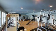 This fitness center offers a thoughtfully designed workout environment spread across a single floor. The space features warm hardwood flooring throughout, complemented by soft grey walls and a smooth ceiling with recessed lighting that provides consistent, even illumination across the entire area. Large wall-mounted mirrors line one side of the room, enhancing the sense of space and allowing for proper form checks during exercise. The cardio section is well-appointed with a variety of machines, including stationary bikes, an elliptical trainer, and a rowing machine, each equipped with digital displays for performance tracking. The strength training area features Quantum cable machines and a squat rack, catering to a full range of resistance-based workouts. A dedicated free weights station with a rack of dumbbells and an adjustable bench rounds out the equipment offerings, supporting both beginners and experienced athletes. The layout is open and organized, allowing for easy movement between stations. The overall ambiance is clean, functional, and professional, making it a practical and well-resourced space for a complete fitness routine.