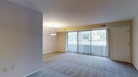 This single-floor space offers a clean, functional layout centered around a generously sized open living area. The room is finished with wall-to-wall carpeting that adds warmth and comfort underfoot, complemented by neutral-toned walls that create a bright, versatile backdrop. A standout feature of the living area is the expansive floor-to-ceiling sliding glass door system fitted with vertical blinds, which spans the full width of the exterior wall. This dramatic glazing allows abundant natural light to pour into the space while offering views of the surrounding outdoor environment, including mature trees visible through the glass. A pendant light fixture hangs overhead, providing focused ambient lighting and serving as a subtle focal point. The ceiling features a textured finish, and HVAC vents are integrated throughout, ensuring climate comfort year-round. The layout includes a defined transitional zone that separates the main living area from adjacent spaces, hinting at a thoughtful and practical floor plan. Overall, the space presents a bright, open ambiance well-suited for comfortable everyday living.