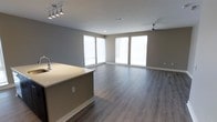 This single-floor space showcases a clean, modern design centered around an open-concept floor plan that connects the kitchen and living areas. The kitchen features a sleek island with a light-toned countertop and an integrated sink, complemented by a track lighting fixture overhead that provides focused, adjustable illumination. The living area is anchored by a ceiling fan, offering both comfort and functionality. Large windows along the far wall flood the space with natural light, creating a bright and airy atmosphere throughout. The walls are finished in a neutral gray tone, providing a versatile backdrop that complements the gray-toned laminate or luxury vinyl plank flooring, which runs consistently throughout the open area. White baseboards provide a crisp contrast against the flooring and walls, adding a polished finish to the overall aesthetic. The spacious, unobstructed layout of the living area allows for flexible furniture arrangements, making the space highly adaptable. Together, these elements create a cohesive, contemporary environment that feels both functional and inviting.