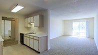 This single-floor residence offers a clean and functional layout designed for comfortable everyday living. The kitchen is positioned at the heart of the space, featuring white cabinetry with ample upper and lower storage, a single-basin sink with a countertop workspace, and a built-in appliance bay for a dishwasher. An under-cabinet light fixture provides focused task lighting, while a skylight above adds a natural brightness to the cooking area. The kitchen flows seamlessly into the adjacent living area, which is finished with wall-to-wall carpeting in a neutral tone, creating a warm and cohesive feel throughout. Smooth, light-colored walls throughout the space contribute to a bright and airy ambiance. A textured ceiling runs consistently across both areas, adding subtle visual character. At the far end of the living area, a large sliding or hinged glass door allows natural light to pour in and provides direct access to an outdoor area, enhancing the sense of openness. The straightforward, open-plan design ensures easy movement between the kitchen and living spaces, making the layout both practical and inviting for daily use.