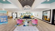 The Sagamore clubhouse is a thoughtfully designed, single-story community space that makes an immediate impression with its dramatic barrel-vaulted ceiling in the central lobby, finished in a dark charcoal tone and anchored by a sculptural globe pendant light fixture. The "Sagamore" script signage mounted high on the white feature wall reinforces the space's strong visual identity. Light wood-look flooring runs throughout, providing a warm, cohesive foundation for the bold color palette. The main living area is furnished with vibrant pink velvet barrel chairs accented with gold frames, a sleek navy sofa with teal and yellow throw pillows, and a vivid geometric area rug, creating a dynamic and inviting lounge environment. A secondary area features striking teal-painted ceiling sections that contrast crisply with white walls, along with black-framed French doors that welcome natural light and outdoor views. Built-in window bench seating offers additional gathering spots. An adjoining room showcases a bold chevron-patterned tile accent wall in yellow, white, and teal, paired with a built-in countertop bar, beverage refrigerator, coffee station, and casual seating — ideal for resident amenity use. Colorful framed artwork and lush hanging greenery complete the retro-modern aesthetic throughout this energetic, well-appointed space.