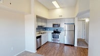 This single-floor space offers a thoughtfully designed layout with a clean, modern aesthetic. The kitchen is a standout feature, boasting stainless steel appliances including a refrigerator, gas range, built-in microwave, and dishwasher. White cabinetry with ample storage lines the walls, complemented by a dual-basin sink and a neutral countertop that provides generous workspace. A two-tone wall treatment — soft white above and a muted blue-gray accent below — adds a subtle design element that gives the kitchen a polished, cohesive look. The soaring ceilings amplify the sense of openness, creating an airy and spacious atmosphere. Warm hardwood-style flooring runs through the kitchen and living areas, providing a welcoming contrast to the bright, light-filled environment. Overhead skylights and recessed ventilation ensure the space remains bright and well-ventilated. The open flow between the kitchen and adjacent living area makes this unit well-suited for everyday living and entertaining alike. Overall, the space strikes a balance between functionality and contemporary design, offering a comfortable and practical living environment.