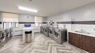 Located at 28 Helene St N in Mississauga, Ontario, this shared laundry facility is a thoughtfully designed, single-floor space that offers a clean and functional environment for residents. The room is finished with a distinctive herringbone-patterned vinyl plank floor in soft grey tones, lending a modern aesthetic throughout.

The walls feature a two-tone tile treatment — crisp white subway tiles on the lower half accented by a bold dark grey tile border — creating a clean, polished look. A row of modern stainless steel front-load washing machines lines one wall, each equipped with digital controls for ease of use. Complementary dryers are positioned nearby, ensuring a complete and efficient laundry experience. A utility sink with a dark wood cabinet provides additional convenience.

Generous white folding counters offer ample workspace for sorting and folding laundry. A seating area with upholstered grey armchairs and a small coffee table allows residents to wait comfortably. Natural light enters through a window fitted with a roller shade, while overhead fluorescent fixtures ensure bright, even illumination throughout. A waste bin and wall-mounted soap dispenser add practical finishing touches. The space is well-organized, well-lit, and designed to serve residents efficiently.
