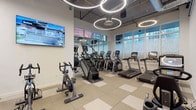 This modern fitness center offers a spacious, single-floor layout designed to accommodate a full range of workout needs. The space is defined by its striking industrial-style aesthetic, featuring exposed concrete columns, open ductwork overhead, and a neutral tiled floor in a checkered pattern that combines durability with visual appeal.

The cardio zone is well-appointed with multiple treadmills, elliptical machines, a stair climber, and both upright and recumbent stationary bikes. Large windows along one wall flood this area with natural light and offer views of the surrounding urban environment, while wall-mounted flat-screen televisions provide entertainment during workouts.

The strength training area is equally comprehensive, featuring a full rack of Matrix-brand dumbbells, adjustable benches, cable machines, and various resistance training stations. Floor-to-ceiling mirrors line the walls, amplifying the sense of space and allowing users to monitor their form.

Throughout the facility, a series of circular LED pendant lights serve as a bold design statement, casting bright, even illumination across the entire space. A water fountain station is conveniently accessible within the gym. The open floor plan ensures ample room to move freely between equipment zones, making this fitness center both functional and visually impressive.