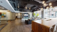 Located at 1222 W Madison St in Chicago's West Loop neighborhood, this single-floor amenity space combines industrial and contemporary design elements to create a sophisticated communal environment. The main lounge area features rich hardwood flooring, an exposed ceiling with dark-painted ductwork, and pendant globe lighting that adds warmth and character. Large street-facing windows flood the seating area with natural light, framing views of the surrounding urban streetscape. Comfortable furnishings — including leather sofas, upholstered wingback chairs, and layered area rugs — are arranged around a built-in media wall with integrated shelving, creating an inviting gathering space. A glass-walled conference room offers a transparent, professional setting equipped with office chairs and display screens, while maintaining visual connectivity to the open lounge. A dedicated meditation room provides a tranquil retreat, finished with deep navy textured walls, a projector, and yoga mats arranged on a tiered platform floor. A kitchenette area rounds out the space, featuring marble-look countertops, light wood cabinetry, a stainless steel sink, and curated artwork. Together, these distinct zones deliver a well-appointed, multi-functional space suited for work, relaxation, and wellness.