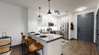 This thoughtfully designed single-floor residence blends contemporary style with functional living. The open-concept kitchen serves as a centerpiece, showcasing white shaker upper cabinets paired with warm-toned lower cabinetry, a classic white subway tile backsplash, and granite countertops. A peninsula island with seating for multiple bar-stool guests creates an inviting space for dining and entertaining. Stainless steel appliances — including a French door refrigerator, electric range, and over-the-range microwave — complete the kitchen with a polished, modern finish. Pendant lighting with matte black accents and a track light fixture illuminate the space with a clean, industrial touch. Wood-look flooring flows throughout, adding warmth and visual continuity.

The bedroom is a standout feature, adorned with a striking hand-drawn mural celebrating the Dallas Farmers Market, complete with botanical illustrations and bold linework. Natural light filters through multiple windows dressed with floor-length gray curtains, creating a cozy yet airy atmosphere.

The bathroom is equally well-appointed, featuring a dual-sink vanity with granite countertops, white shaker cabinetry, matte black hardware, a large framed mirror, and a tiled shower with a curved curtain rod. A walk-in closet with shelving is accessible directly from the bathroom, adding practical storage to this well-curated space.