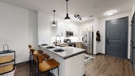 This thoughtfully designed single-floor residence blends contemporary style with functional living. The open-concept kitchen serves as a centerpiece, showcasing white shaker upper cabinets paired with warm-toned lower cabinetry, a classic white subway tile backsplash, and granite countertops. A peninsula island with seating for multiple bar-stool guests creates an inviting space for dining and entertaining. Stainless steel appliances — including a French door refrigerator, electric range, and over-the-range microwave — complete the kitchen with a polished, modern finish. Pendant lighting with matte black accents and a track light fixture illuminate the space with a clean, industrial touch. Wood-look flooring flows throughout, adding warmth and visual continuity.

The bedroom is a standout feature, adorned with a striking hand-drawn mural celebrating the Dallas Farmers Market, complete with botanical illustrations and bold linework. Natural light filters through multiple windows dressed with floor-length gray curtains, creating a cozy yet airy atmosphere.

The bathroom is equally well-appointed, featuring a dual-sink vanity with granite countertops, white shaker cabinetry, matte black hardware, a large framed mirror, and a tiled shower with a curved curtain rod. A walk-in closet with shelving is accessible directly from the bathroom, adding practical storage to this well-curated space.