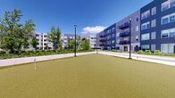 Located at 7244 N Lincoln Ave in Lincolnwood, IL, this thoughtfully designed outdoor courtyard serves as a central amenity space nestled within a contemporary multi-story residential building. The space is anchored by a generously sized artificial turf putting green, complete with flagged holes, offering residents a convenient and enjoyable recreational outlet right outside their door. The lush green surface contrasts beautifully with the surrounding hardscape pathways and manicured garden beds, which feature a variety of shrubs, flowering plants, and mature trees that add color and texture throughout the seasons. A covered pergola structure defines a dedicated outdoor lounge and dining area, furnished with comfortable seating arrangements including chairs, sofas, and a dining table, making it an inviting space for relaxation and social gatherings. Tall, modern lamp posts are positioned throughout the courtyard, ensuring the space remains well-lit and functional into the evening hours. The surrounding building features a contemporary exterior with gray and blue paneling, large windows, and private balconies that overlook the courtyard below. The entire space benefits from abundant natural light under open skies, creating a bright and welcoming atmosphere. This well-appointed outdoor amenity area reflects a strong commitment to resident lifestyle and community living.