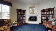 Located at 232 W. Gilbert St. in Hampton, Virginia, this single-floor space exudes a classic, refined character throughout. The standout feature is a beautifully appointed library room, designed to offer both comfort and sophistication. A corner fireplace serves as the room's focal point, framed by a clean white mantel adorned with decorative accents and a framed landscape painting above. Flanking the fireplace are two stately mahogany bookcases with arched glass-panel doors, filled with an extensive collection of books — lending the space an atmosphere of scholarly elegance. Rich crown molding lines the ceiling, adding architectural detail that enhances the room's traditional aesthetic. The deep blue carpeting grounds the space with a sense of warmth, complementing the neutral wall tones and dark wood furnishings. A delicate side table positioned near the bookcases provides a thoughtful reading nook. The overall ambiance is one of quiet sophistication — ideal for reading, reflection, or intimate conversation. The careful attention to traditional design elements, from the millwork to the curated furnishings, makes this a space that feels both purposeful and inviting.