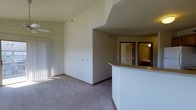 This single-floor space is characterized by its vaulted ceiling, which rises dramatically above the main living area to create an expansive, airy feel. The open layout is anchored by a curved breakfast bar with a warm-toned countertop, serving as a natural divider between the living space and the adjacent area beyond. Neutral-toned carpeting runs throughout, complementing the light gray walls and white baseboards for a clean, cohesive look. Natural light filters into the living area through vertical blinds on what appears to be a sliding glass door, brightening the space. Toward the rear, a hallway leads to additional rooms, with wood-framed doors visible and a lit interior space suggesting a bathroom or secondary room. The overall design balances openness with functional separation, making efficient use of the single-floor footprint. The vaulted ceiling remains the standout architectural element, adding volume and visual interest to an otherwise straightforward and practical layout suited for comfortable everyday living.
