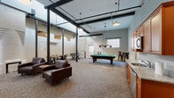 This single-floor community lounge showcases a striking industrial aesthetic rooted in historic architecture. Painted white brick walls, exposed structural steel columns, and soaring ceilings with black steel beams create a raw yet refined atmosphere throughout the space. Abundant natural light filters in through large arched and grid-pane windows, brightening the open layout and highlighting the textured carpet flooring beneath.

The main gathering area is anchored by a classic pool table illuminated by a trio of pendant lights, offering a central recreational focal point. Nearby, a comfortable lounge zone features rich brown leather armchairs arranged around a small side table, with a wall-mounted flat-screen TV providing an entertainment hub. The seating area is further complemented by a foosball table and additional bistro-style seating, catering to a variety of social activities.

A well-appointed kitchenette along one wall includes warm wood cabinetry, granite countertops, a stainless steel sink, dishwasher, and microwave—ideal for gathering refreshments. An arched brick alcove adds a charming architectural detail near the kitchenette. The overall ambiance is energetic yet inviting, balancing the character of a repurposed historic building with comfortable, modern furnishings suited for communal use.
