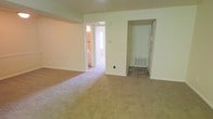 This single-floor residential space offers a clean, functional layout designed for comfortable everyday living. The main living area is finished with wall-to-wall carpeting in a neutral beige tone, providing a warm and quiet underfoot feel throughout. Soft, cream-colored walls create a bright and airy atmosphere, complementing the overall simplicity of the interior design. The space features an open passage leading into an adjoining room, which appears to be a bathroom or utility area with natural light filtering through a window, adding depth and connectivity to the floor plan. A separate doorway leads to an additional room, likely a bedroom or entryway, complete with a built-in HVAC floor vent and an exterior-facing door, suggesting convenient access. The layout benefits from a straightforward flow between spaces, making it easy to navigate. The neutral color palette and carpeted floors provide a versatile canvas suitable for a variety of furnishing styles. Overall, the space presents a tidy, no-frills interior that prioritizes practicality and comfort in a well-connected single-level arrangement.